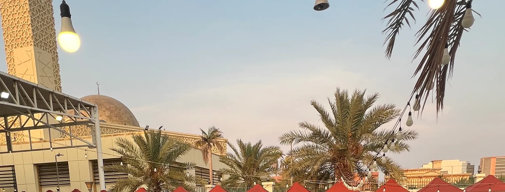 فناء مركز حي المحمدية وقت الغروب، أكشاك حمراء تحت النخيل وسلاسل إنارة، وخلفها مئذنة المسجد وقبته