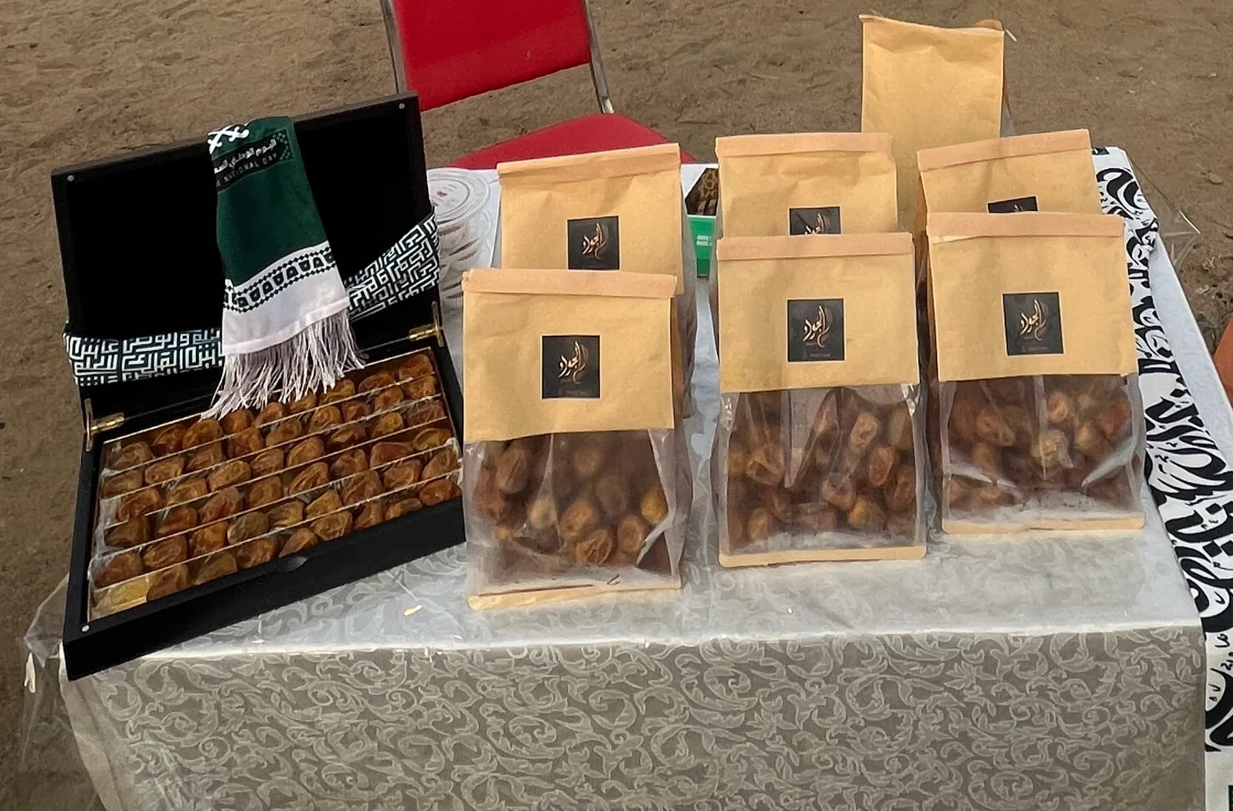 طاولة الجود للتمور الفاخرة في الفعالية: صندوق تمر مفتوح عليه وشاح اليوم الوطني، وستة أكياس ورقية بشعار الجود ونافذة شفافة تبين التمر